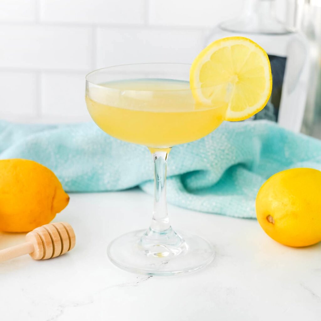 Yellow liquid in a coupe glass with a lemon slice on the rim, a honey dripper, a lemon, and a light blue dishtowel in the background on a white counter.
