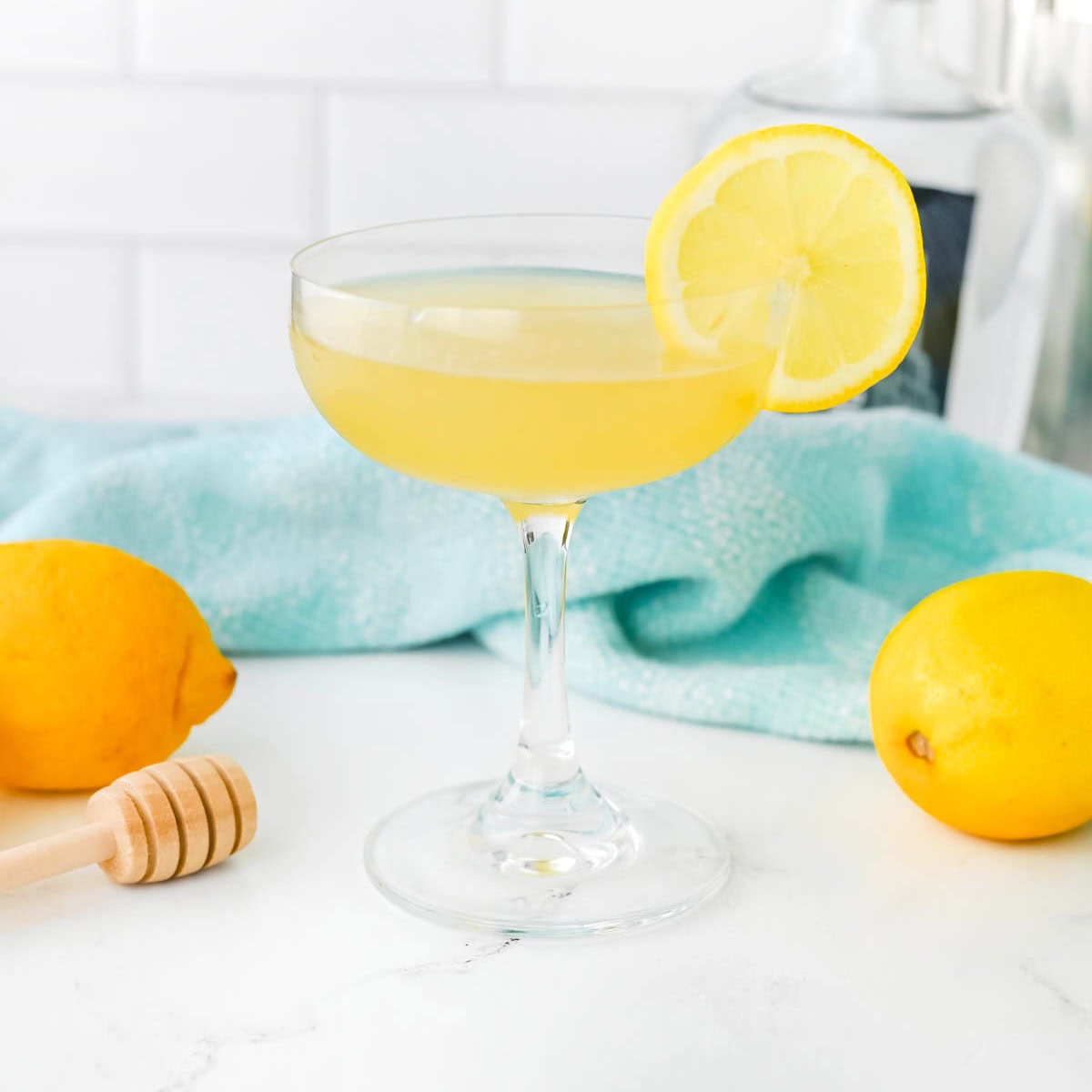 Yellow liquid in a coupe glass with a lemon slice on the rim, a honey dripper, a lemon, and a light blue dishtowel in the background on a white counter.