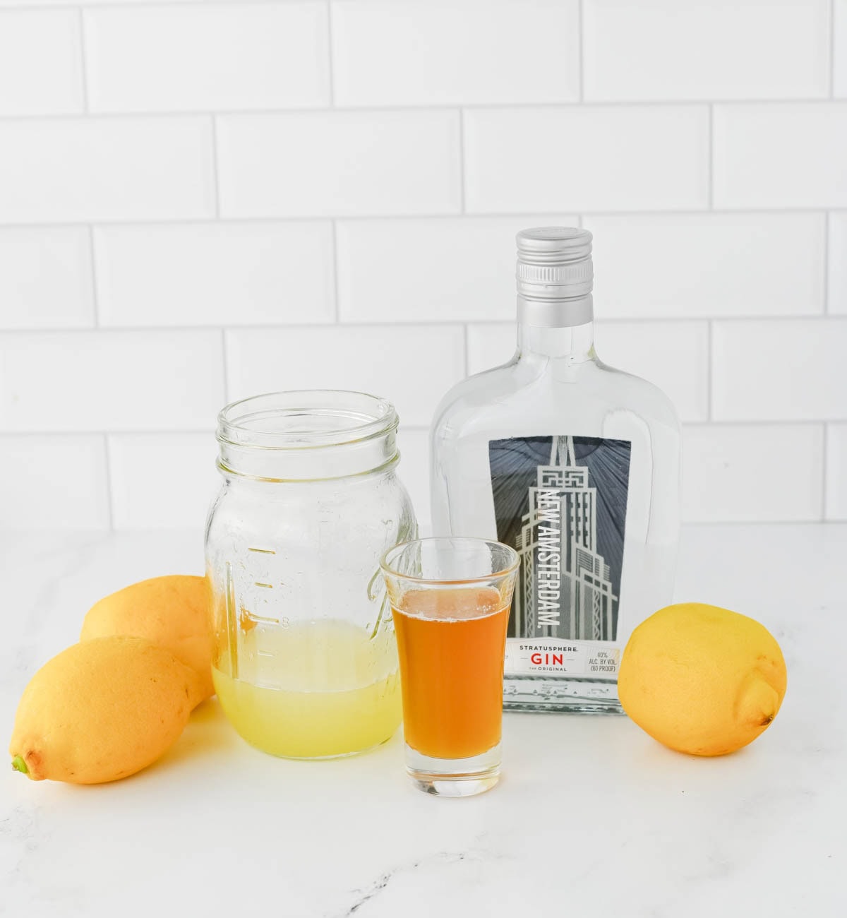 Lemons, a mason jar of lemon juice, a shot glass of honey syrup, and a bottle of gin on a white counter.
