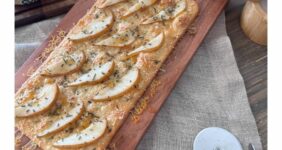 Flatbread with cheese, pears, rosemary, and hot honey on wood board with block of cheese.