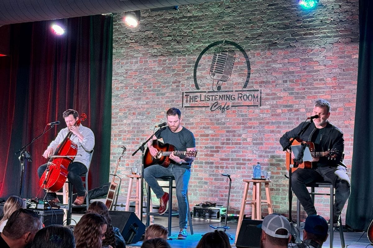 Three men on stage playing insturments with people sitting in audience.