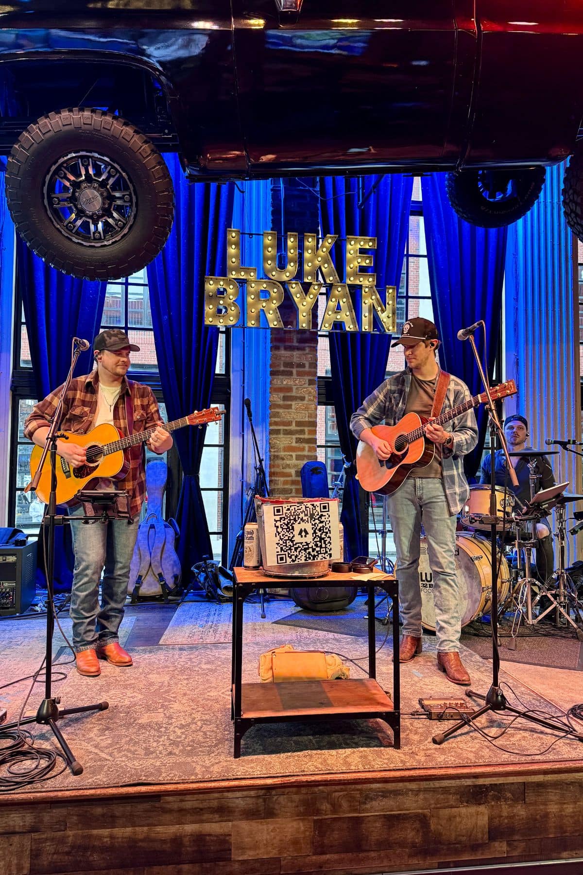 Band playing on a stage with sign that says Luke Bryan and a car hanging overhead.