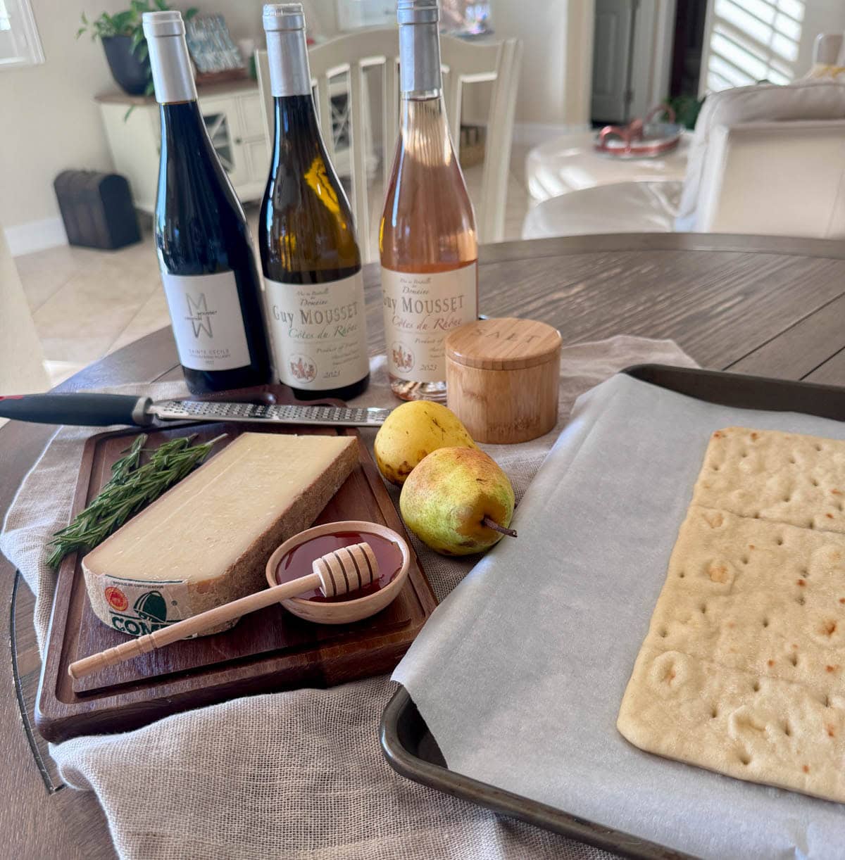 Flatbread on parchment on baking sheet with block of cheese, rosemary, and dish of honey on wood board with salt container and wine in background.