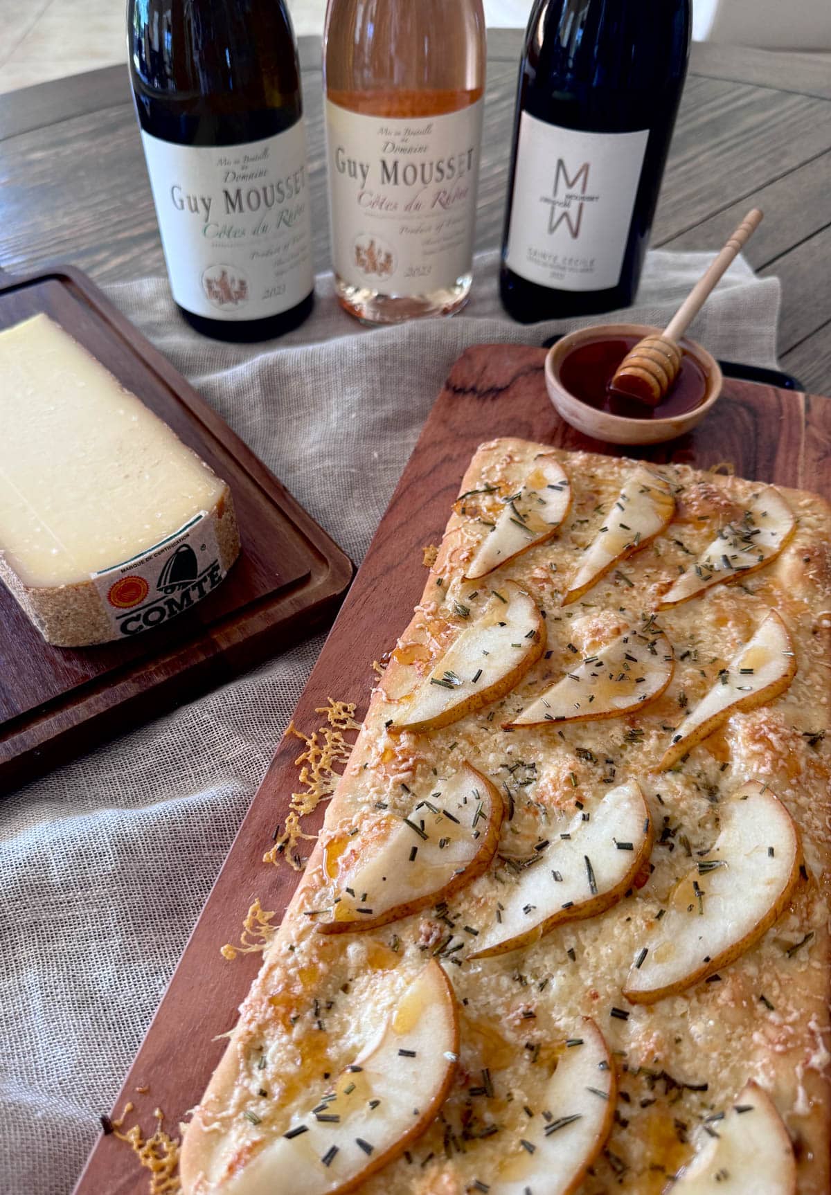 Flatbread with cheese, pears, rosemary, and hot honey on wood board with block of cheese and bottles of wine to the side.