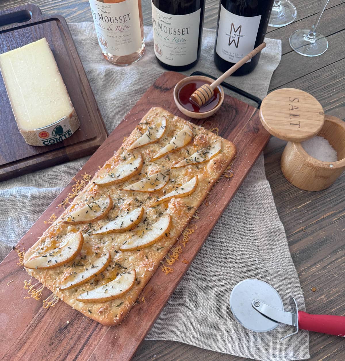 Flatbread with cheese, pears, rosemary, and hot honey on wood board with block of cheese and bottles of wine to the side.
