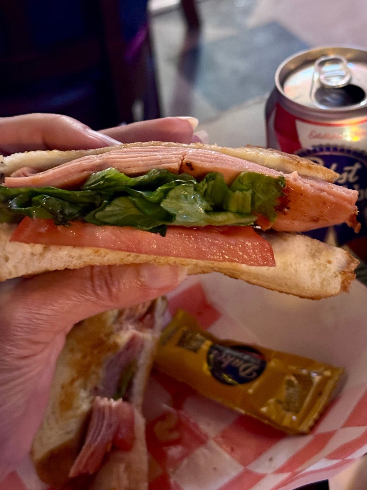Hand holding sandwich of meat, lettuce, and tomato on white bread with can of beer in background.