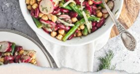 White bowl filled with three kinds of beans, onion, dill, radishes, and dressing, on a wood cutting board on a gray counter.
