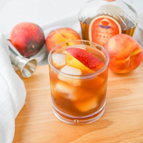 A glass of peach tea with a slice of peach on a cutting board.
