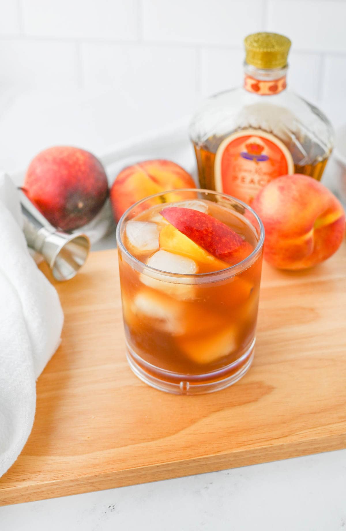 A glass of peach tea with a slice of peach on a cutting board with peaches in background with a whiskey bottle.