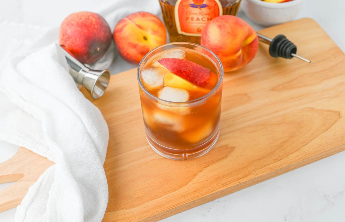 A glass of peach tea with a slice of peach on a cutting board with peaches in background with a whiskey bottle.