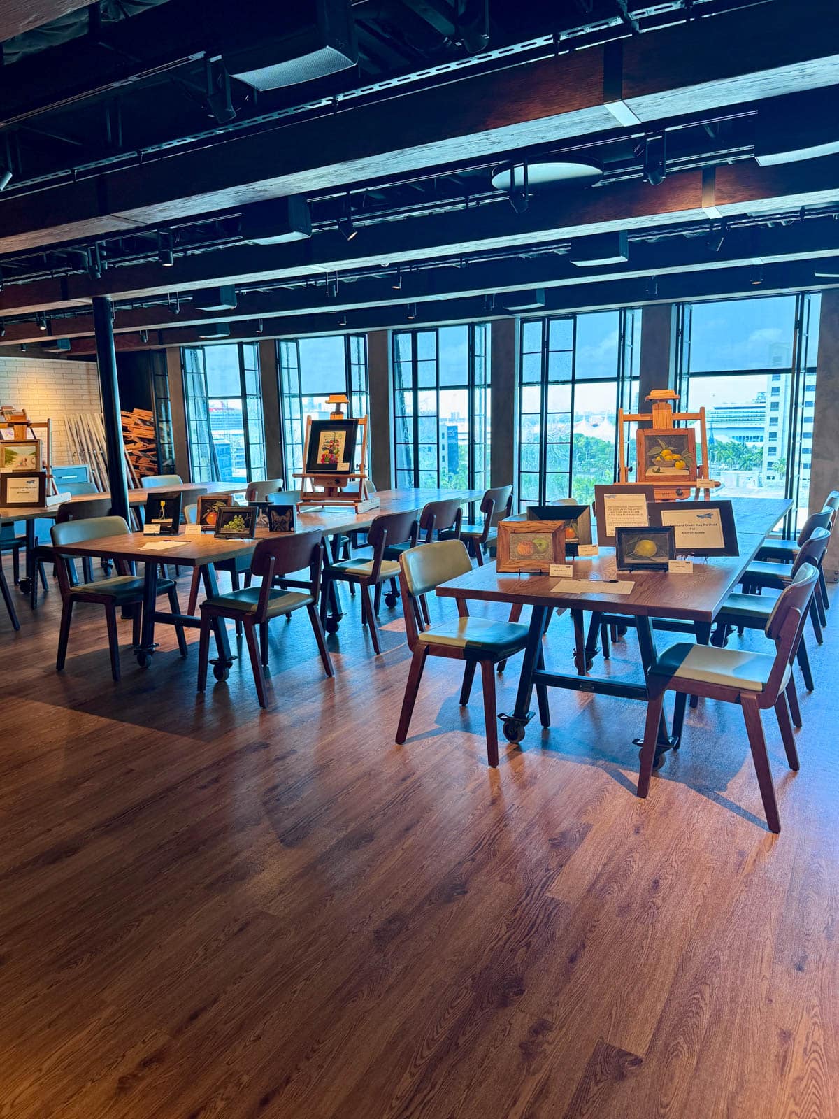 Room with tables and chairs set up for art class with easels and framed paintings.