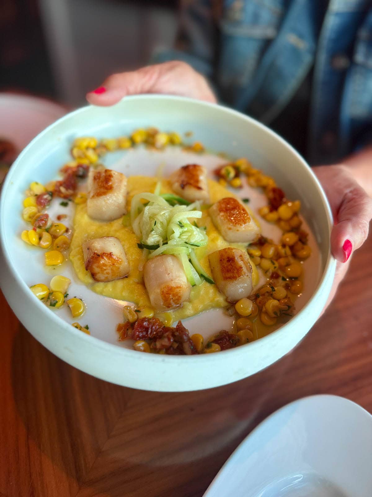 Hands holding a white bowl with scallops and vegetables with sauce.
