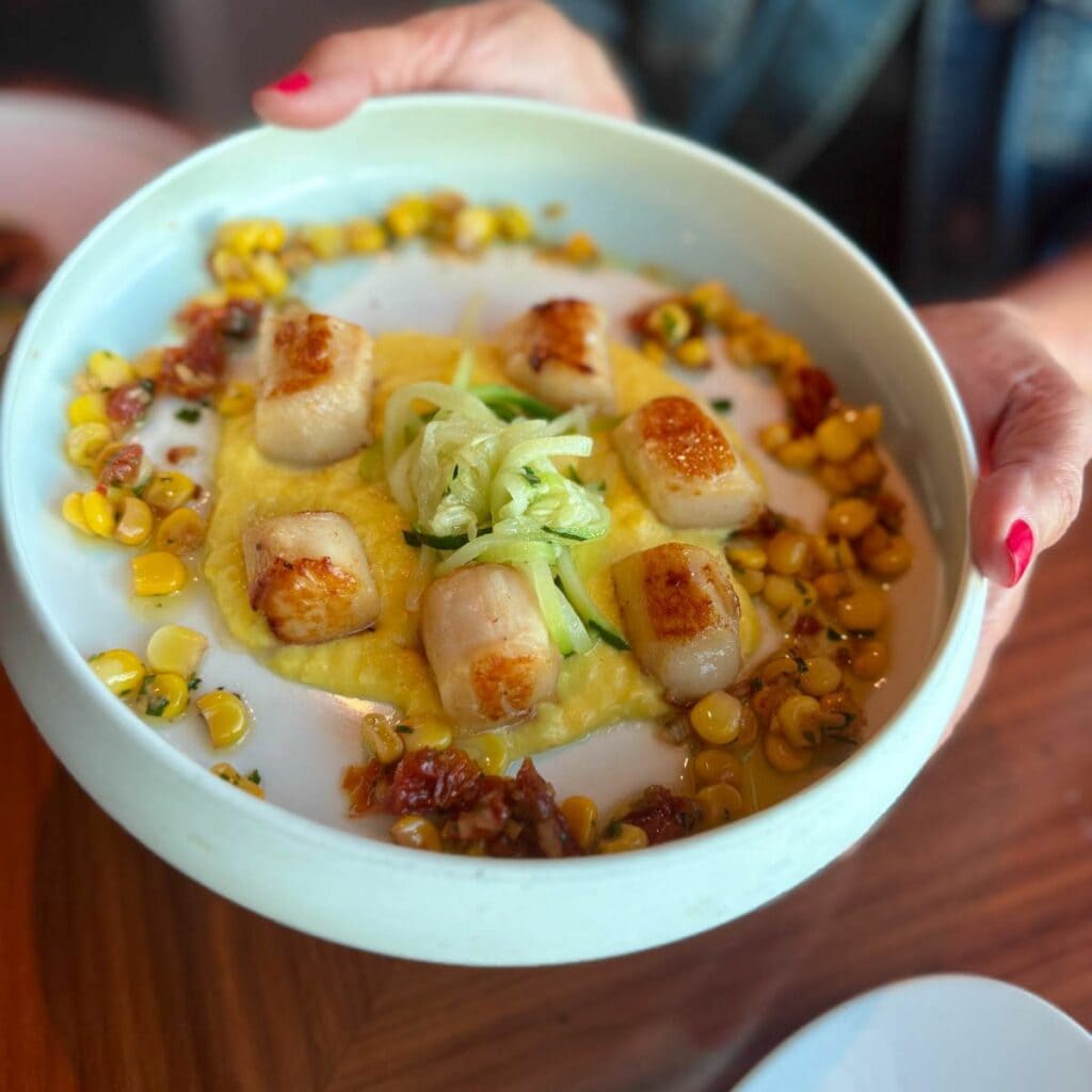 Hands holding a white bowl of scallops with vegetables and sauce.