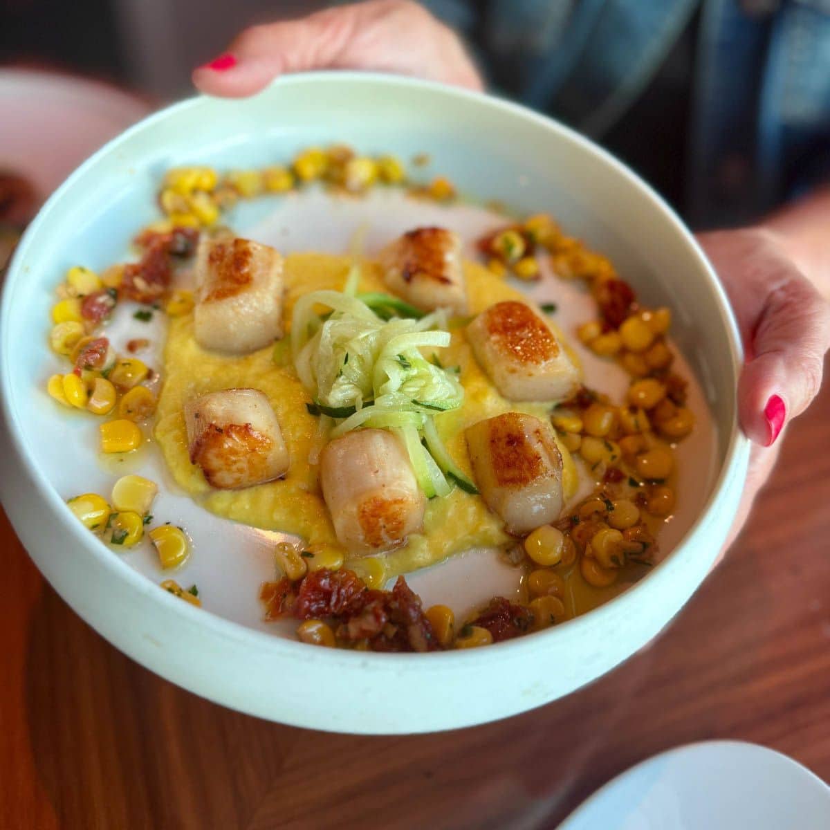 Hands holding a white bowl of scallops with vegetables and sauce.