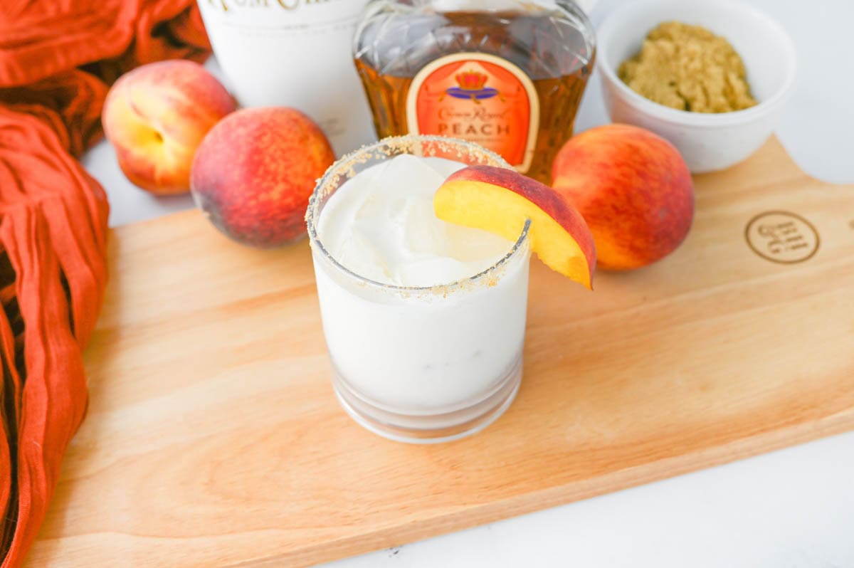 White liquid in a glass with sugar rim on a wood board with peaches and Crown Royal bottle in background.