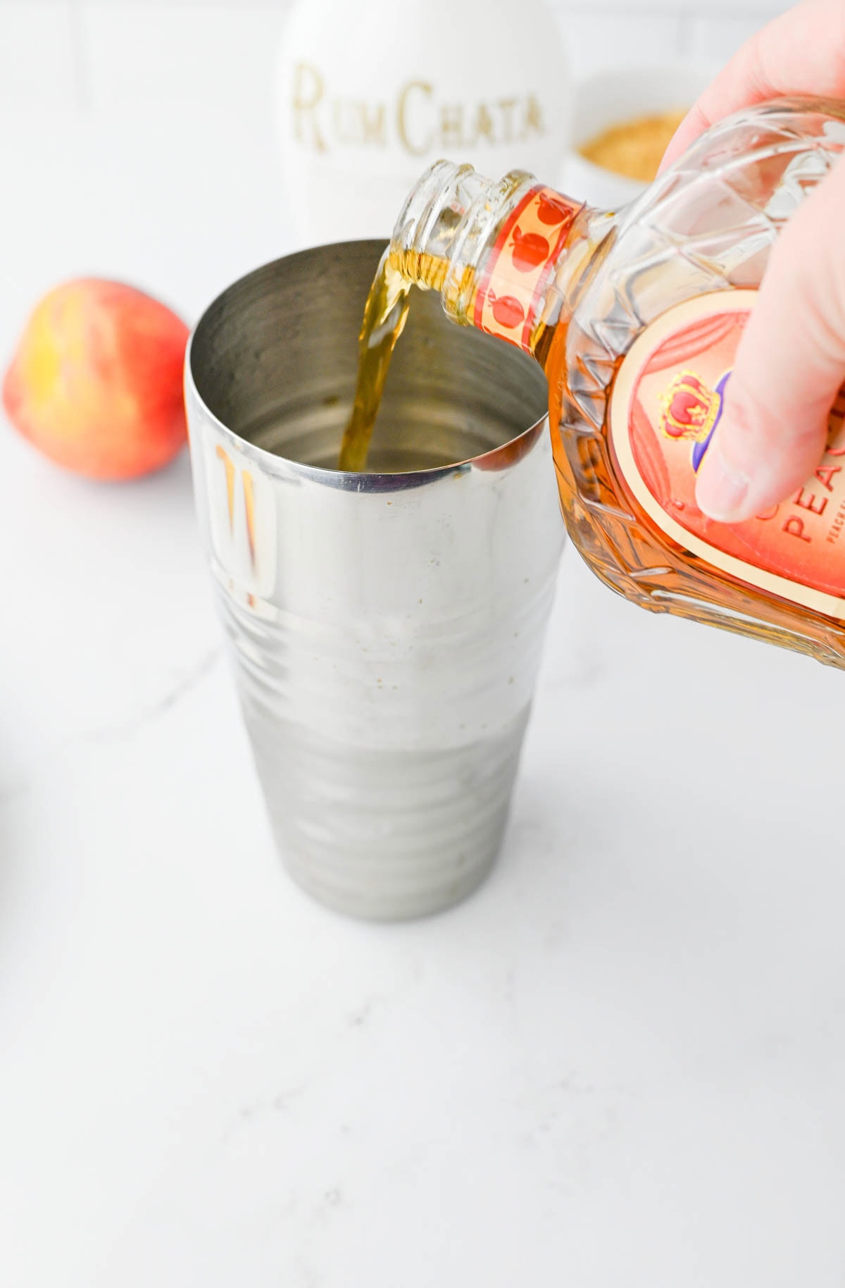 Pouring Crown Royal into a cocktail shaker with Rumchate and peach in background.