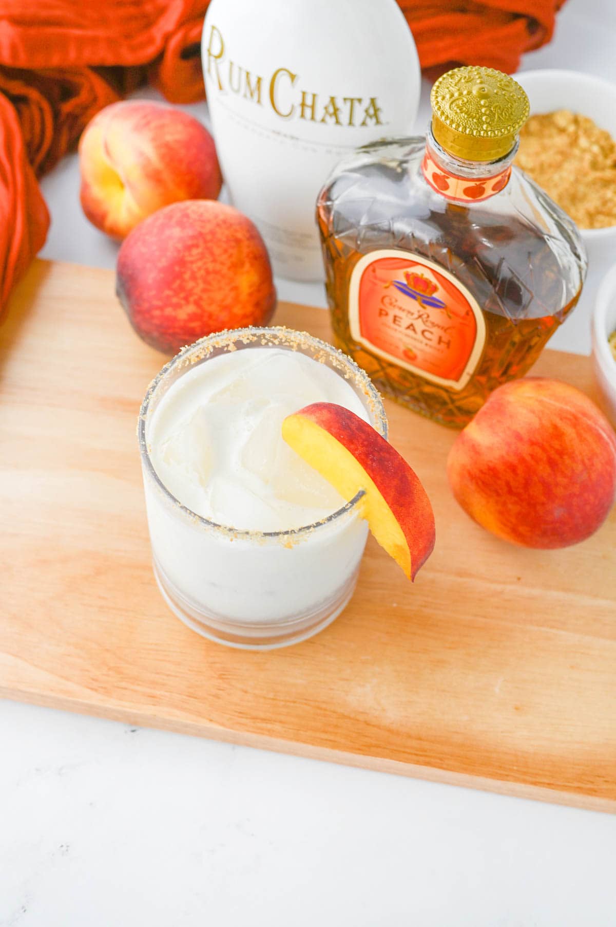 White liquid in a glass with sugar rim on a wood board with peaches and Crown Royal bottle in background.