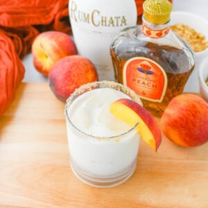 Glass with white liquid and peach slice on wood table with peaches and Crown Royal and Rumchata bottle.