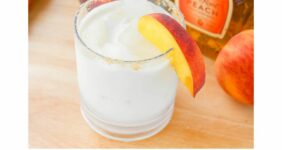 Glass with white liquid and peach slice on wood table with peaches and Crown Royal and Rumchata bottle.