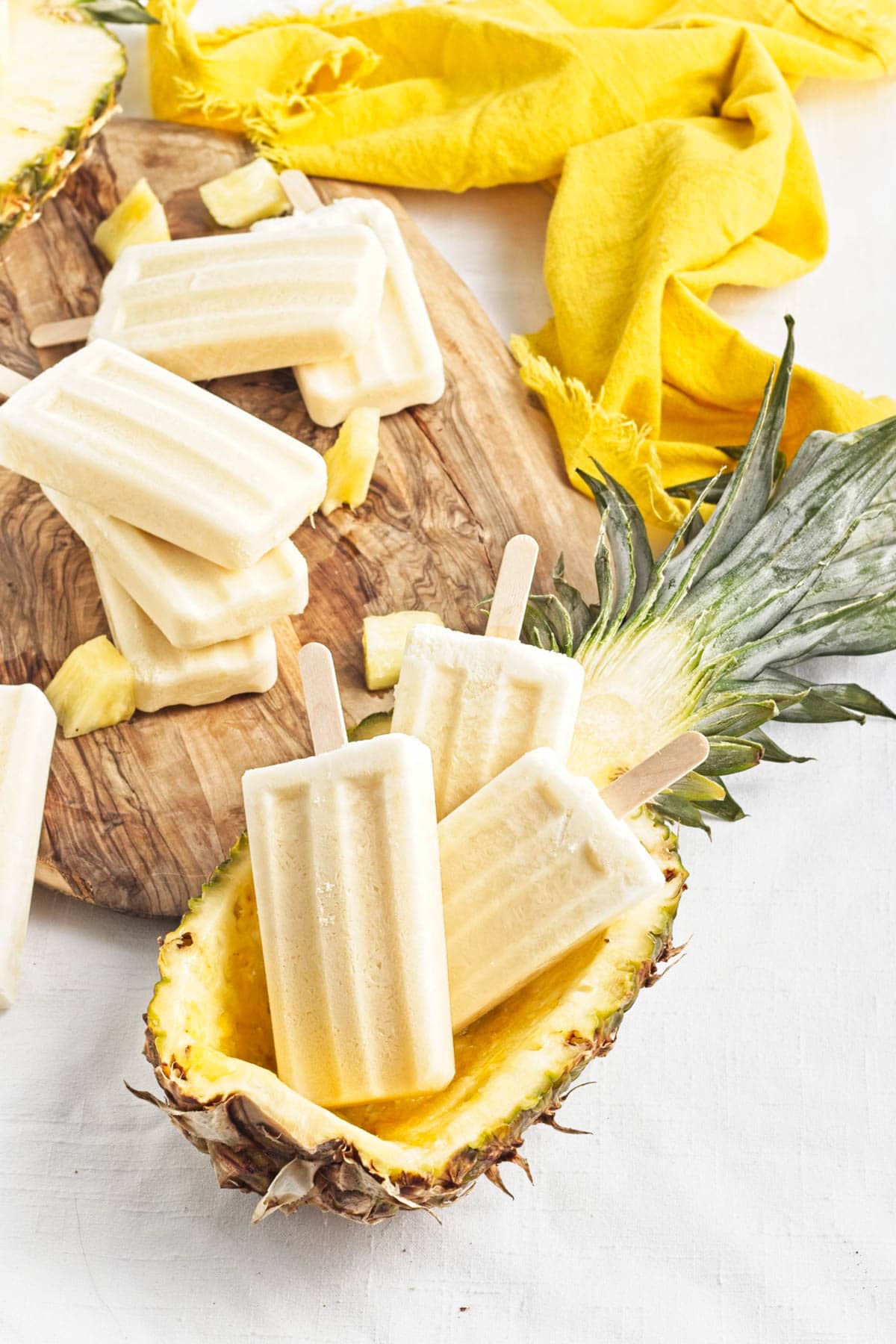 Yellow popsicles on a brown wood board with pineapple chunks, a half pineapple with more popsicles inside, and yellow napkin to the side.