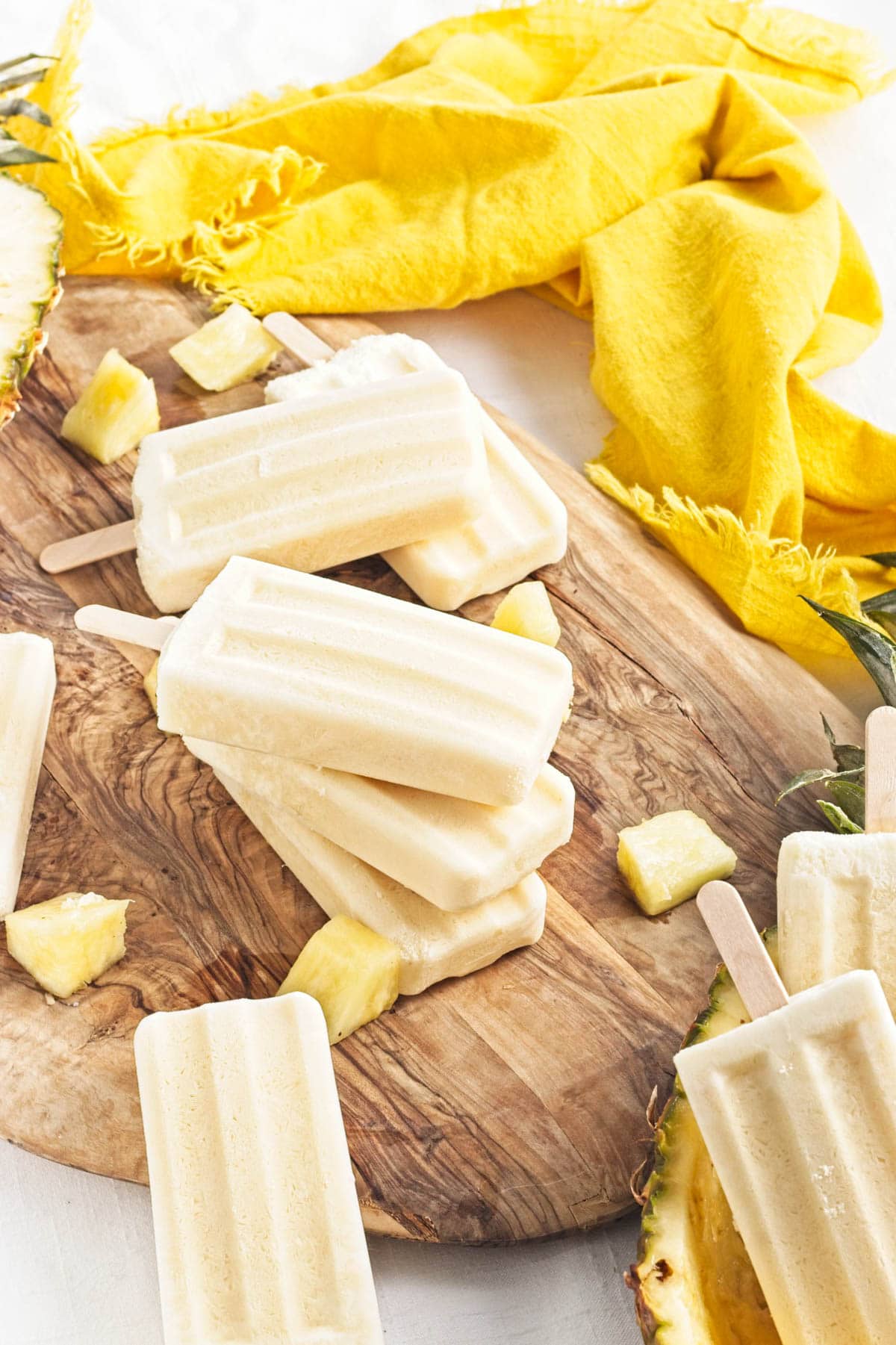Yellow popsicles on a brown wood board with pineapple chunks, a half pineapple, and yellow napkin to the side.