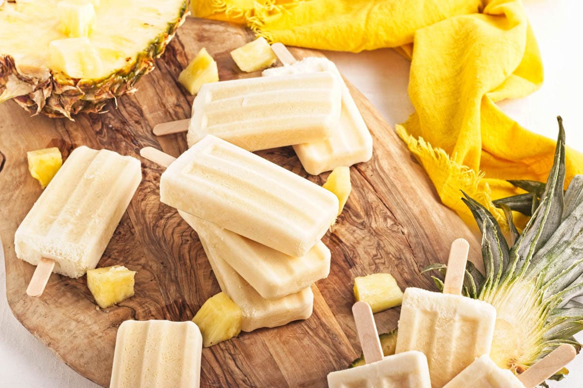 Yellow popsicles on a brown wood board with pineapple chunks, a half pineapple, and yellow napkin to the side.