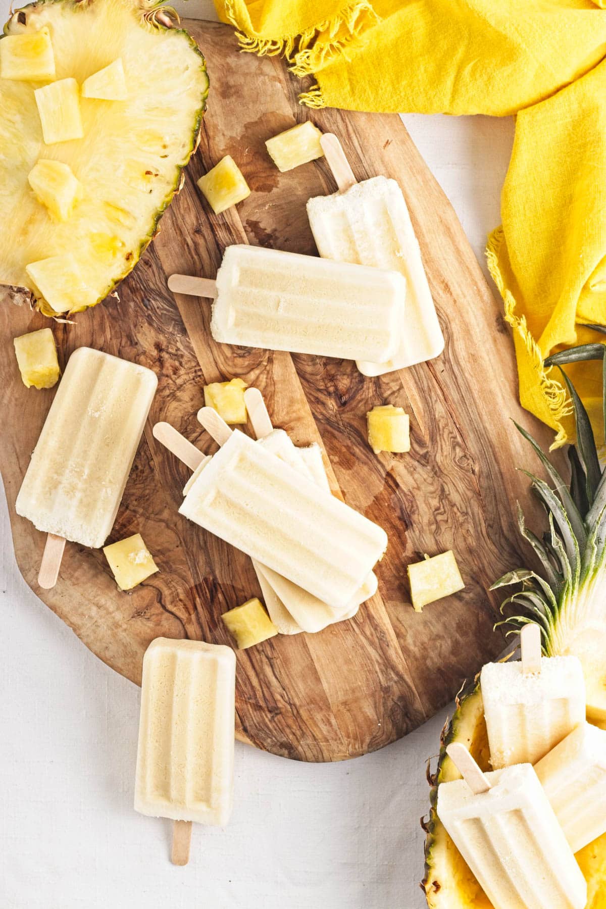 Yellow popsicles on a brown wood board with pineapple chunks, a half pineapple, and yellow napkin to the side.
