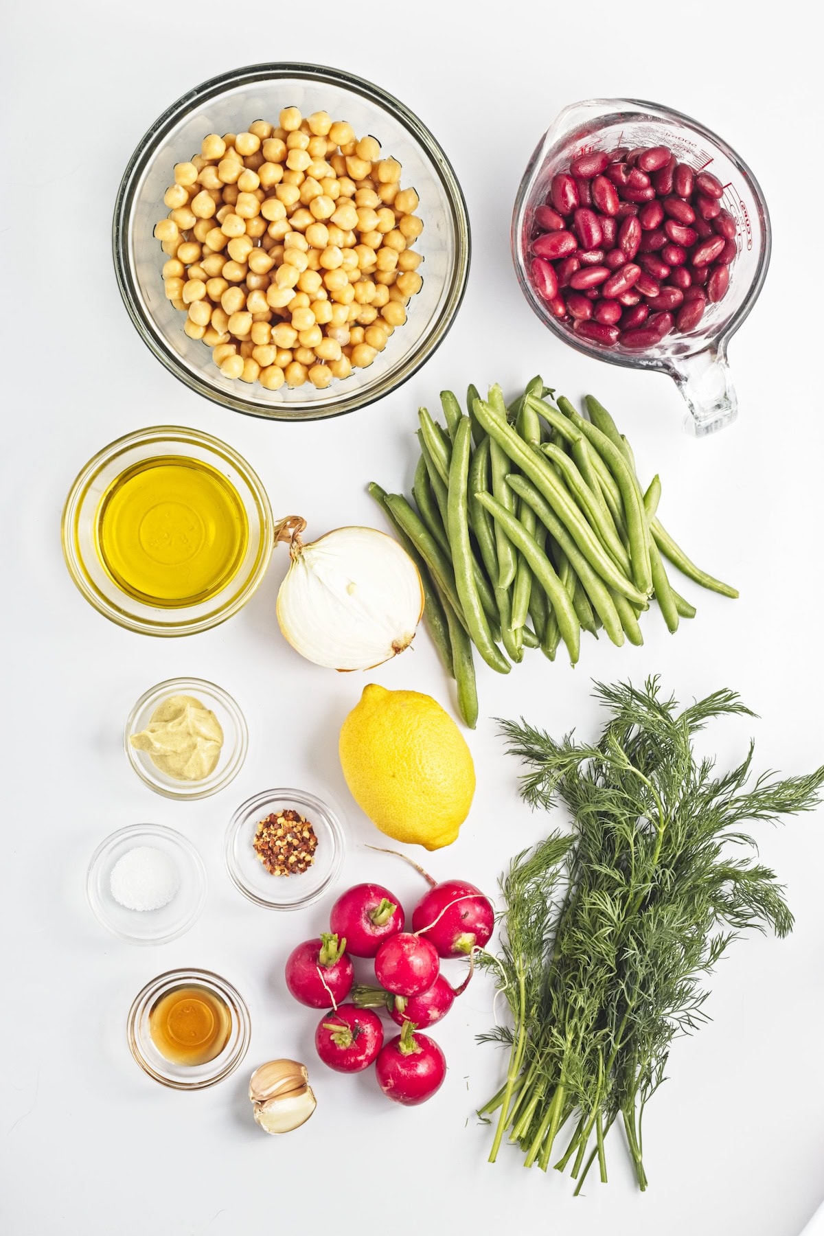 Glass bowls of beans, olive oil, garlic, salt, seasoning, syrup, with bunched of radishes, dill, green beans, and an onion and lemon on counter.