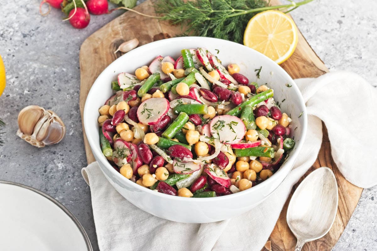 White bowl filled with three kinds of beans, onion, dill, radishes, and dressing, on a wood cutting board on a gray counter.