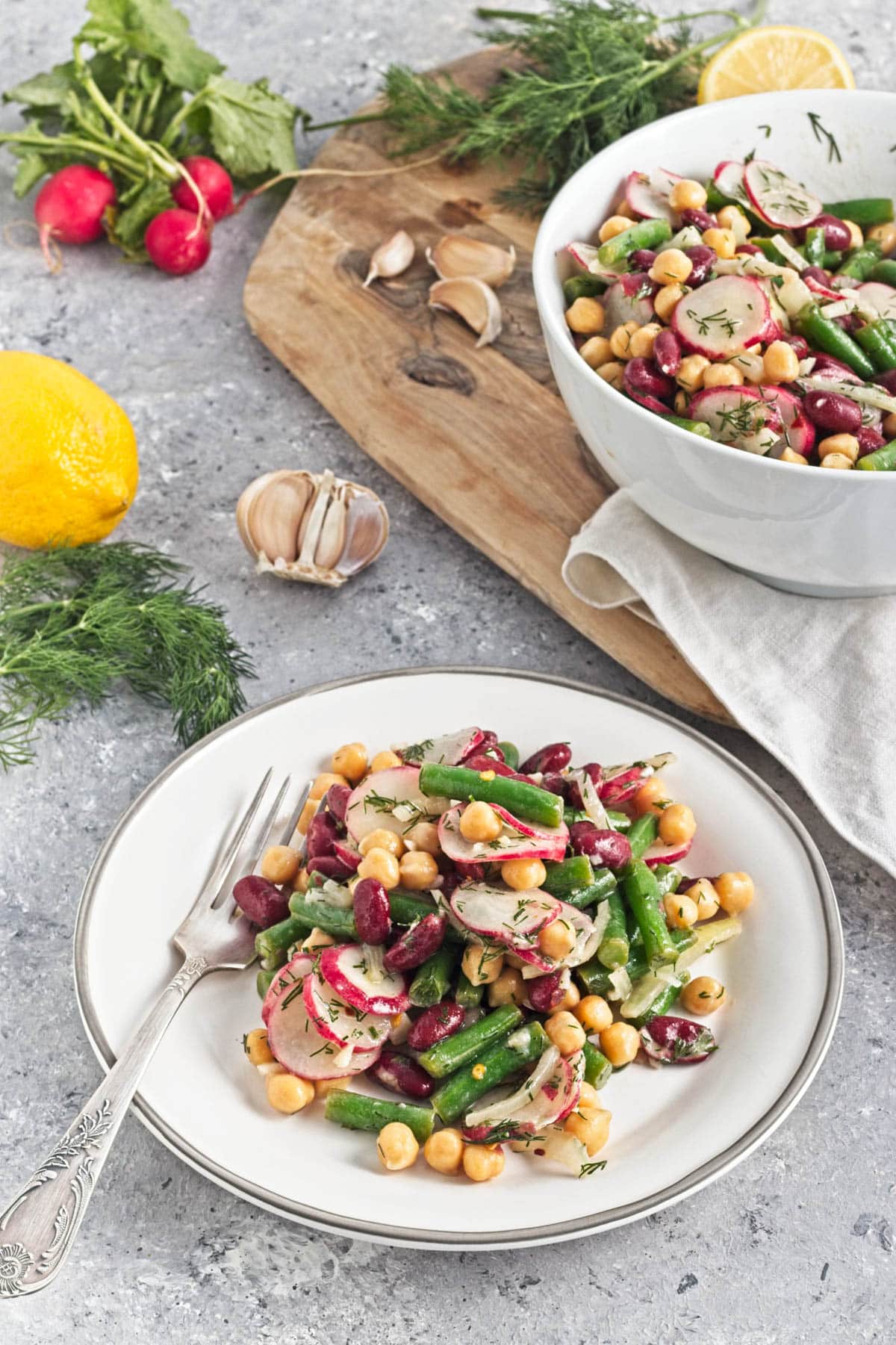 White bowl filled with three kinds of beans, onion, dill, radishes, and dressing, on a wood cutting board.