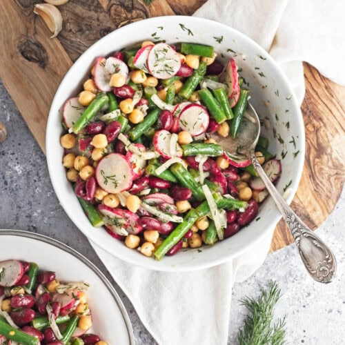 White bowl filled with three kinds of beans, onion, dill, radishes, and dressing, on a wood cutting board.