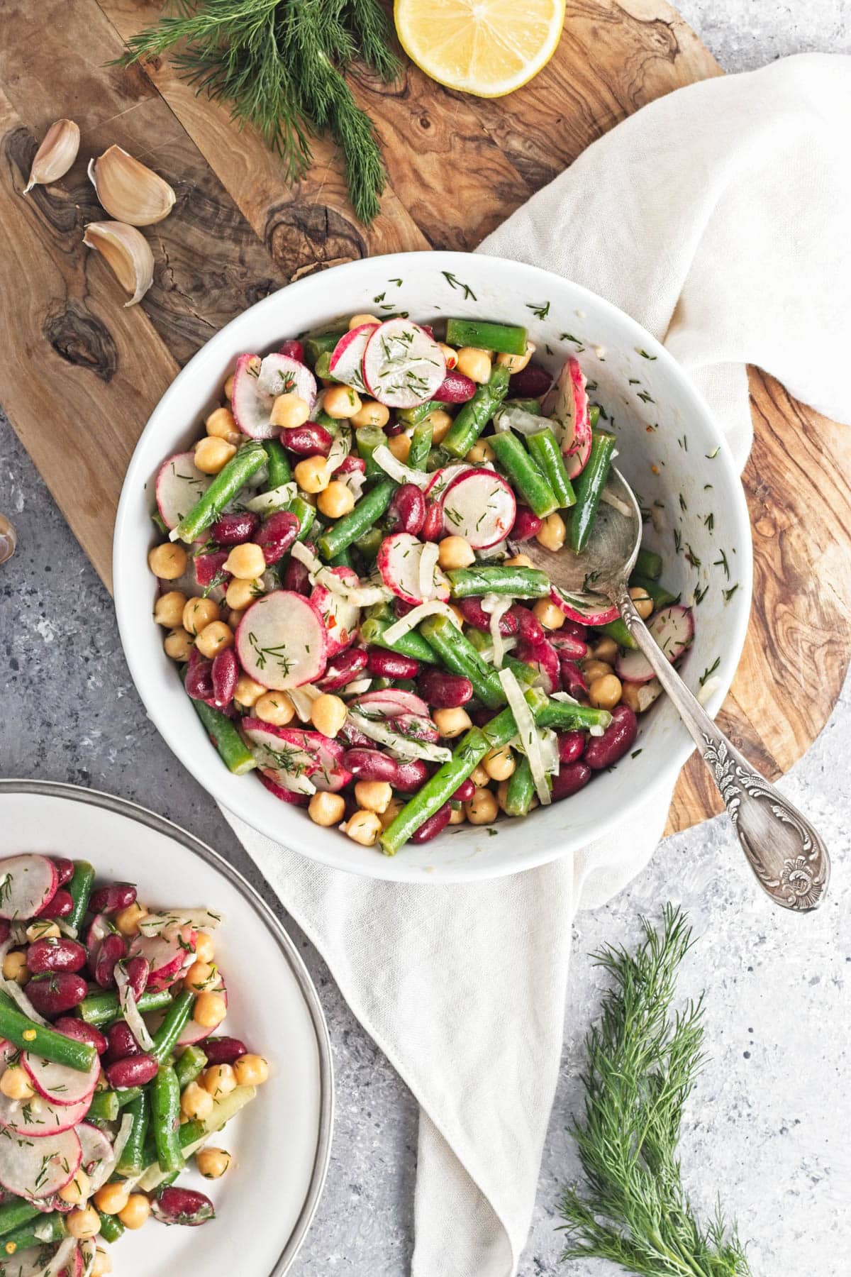 White bowl filled with three kinds of beans, onion, dill, radishes, and dressing, on a wood cutting board.