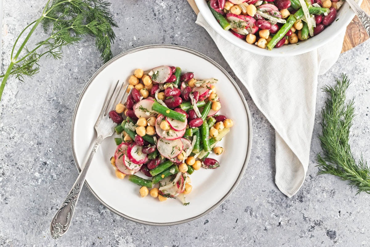 White bowl filled with three kinds of beans, onion, dill, radishes, and dressing, on a wood cutting board.