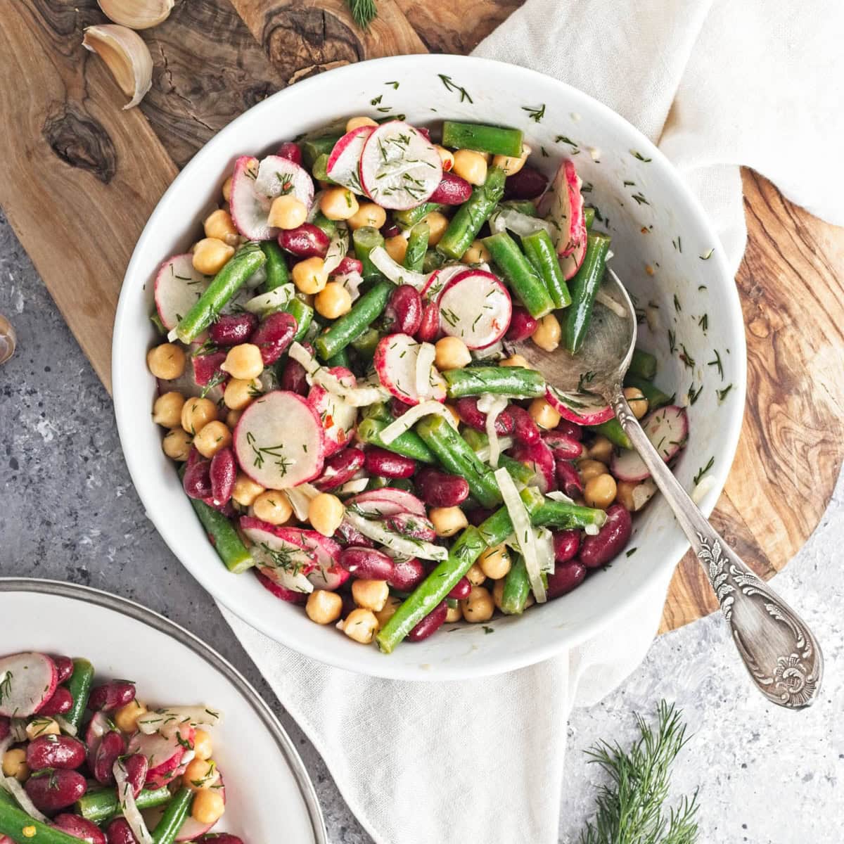 White bowl filled with three kinds of beans, onion, dill, radishes, and dressing, on a wood cutting board on a gray counter.