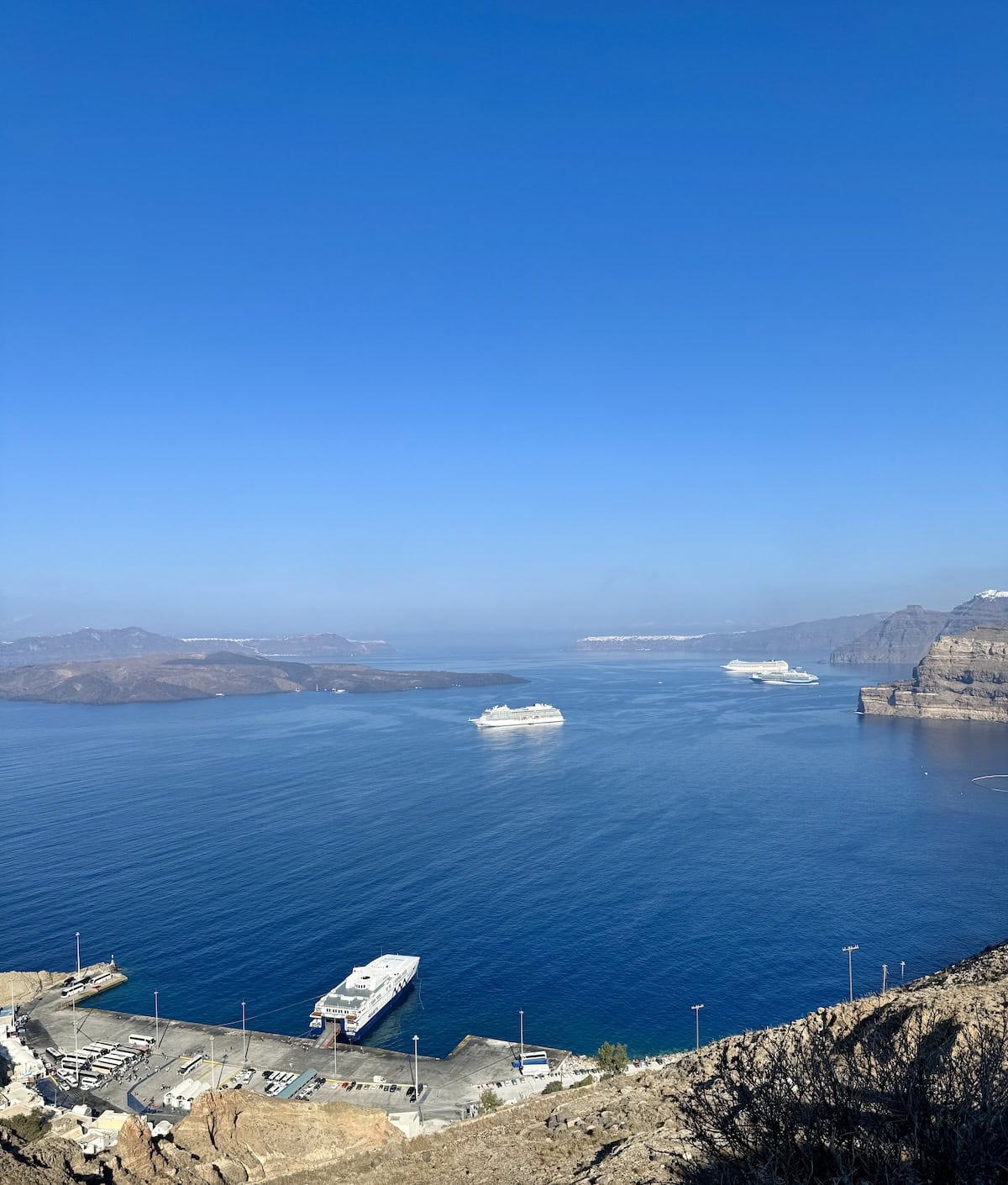 Ferry stop below a cliff with blue sea, blue sky, islands, and cruise ships in the distance. 
