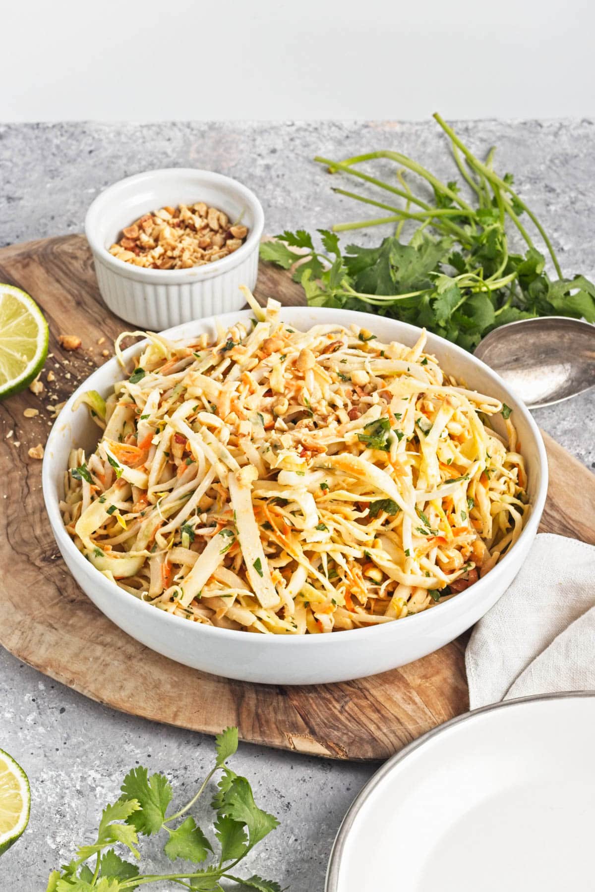A white serving bowl with shredded cabbage mix on a cutting board with limes, cliantro, and a bowl of peanuts.