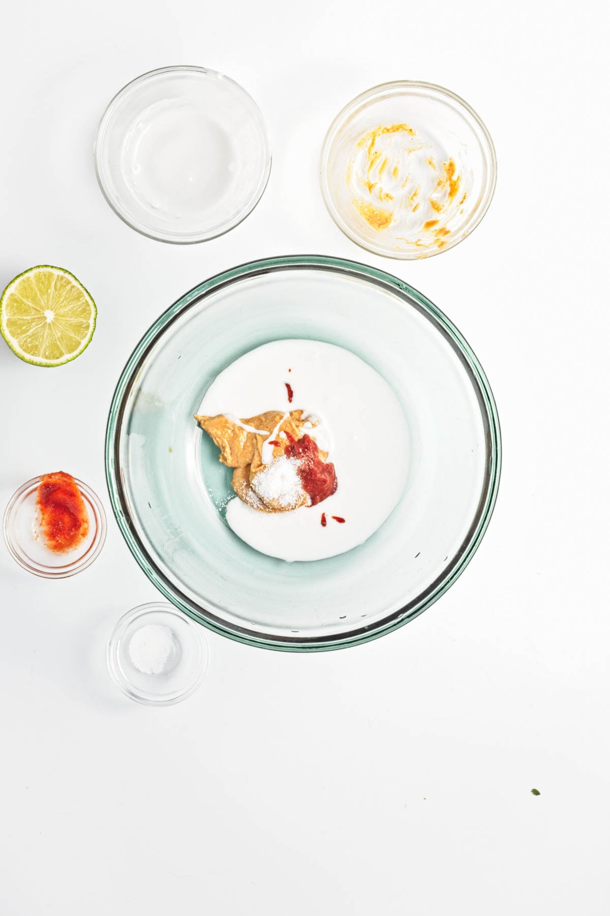 White liquid, salt, red paste, and peanut butter in a glass bowl with empty bowls and a lime half on side.