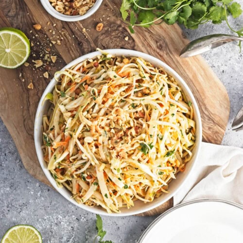 A white serving bowl and plate with shredded cabbage mix on a cutting board with limes, cliantro, and a bowl of peanuts.