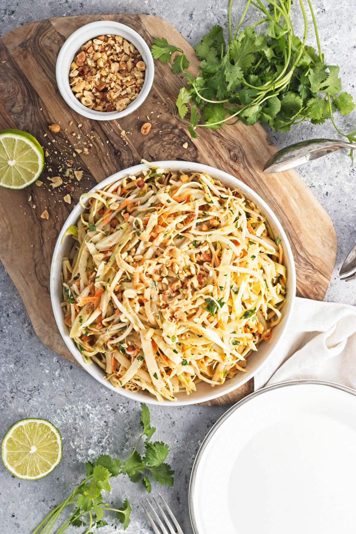 A white serving bowl and plate with shredded cabbage mix on a cutting board with limes, cliantro, and a bowl of peanuts.