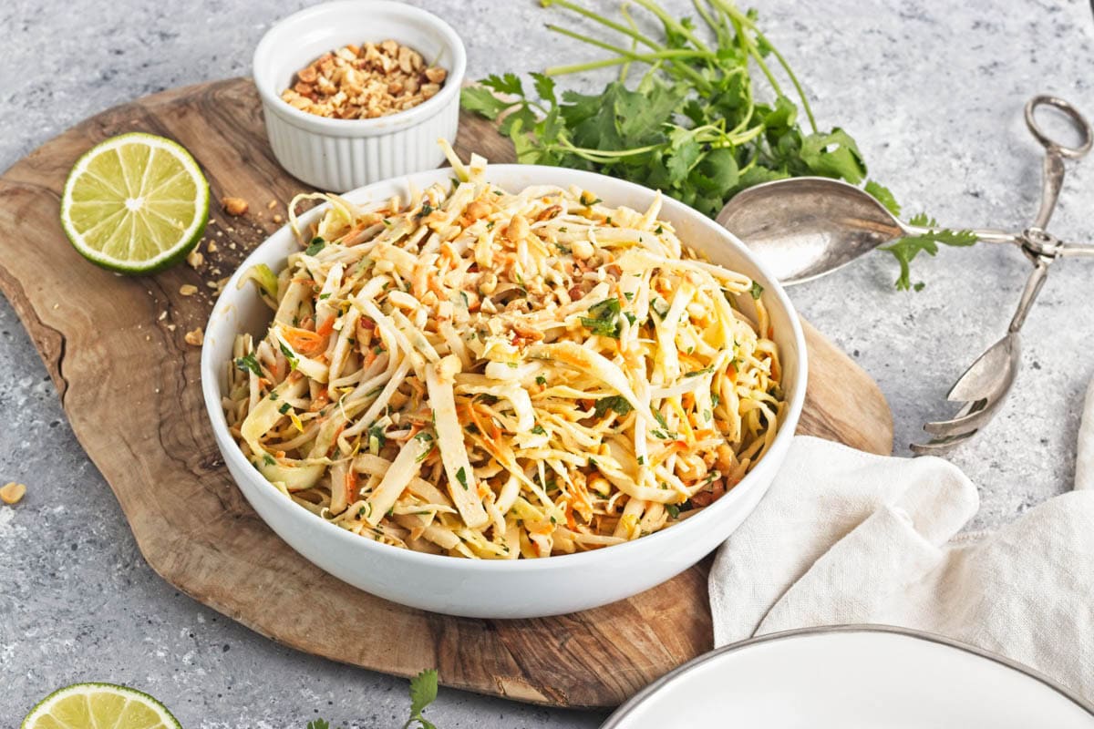 A white serving bowl with shredded cabbage mix on a cutting board with limes, cliantro, and a bowl of peanuts.