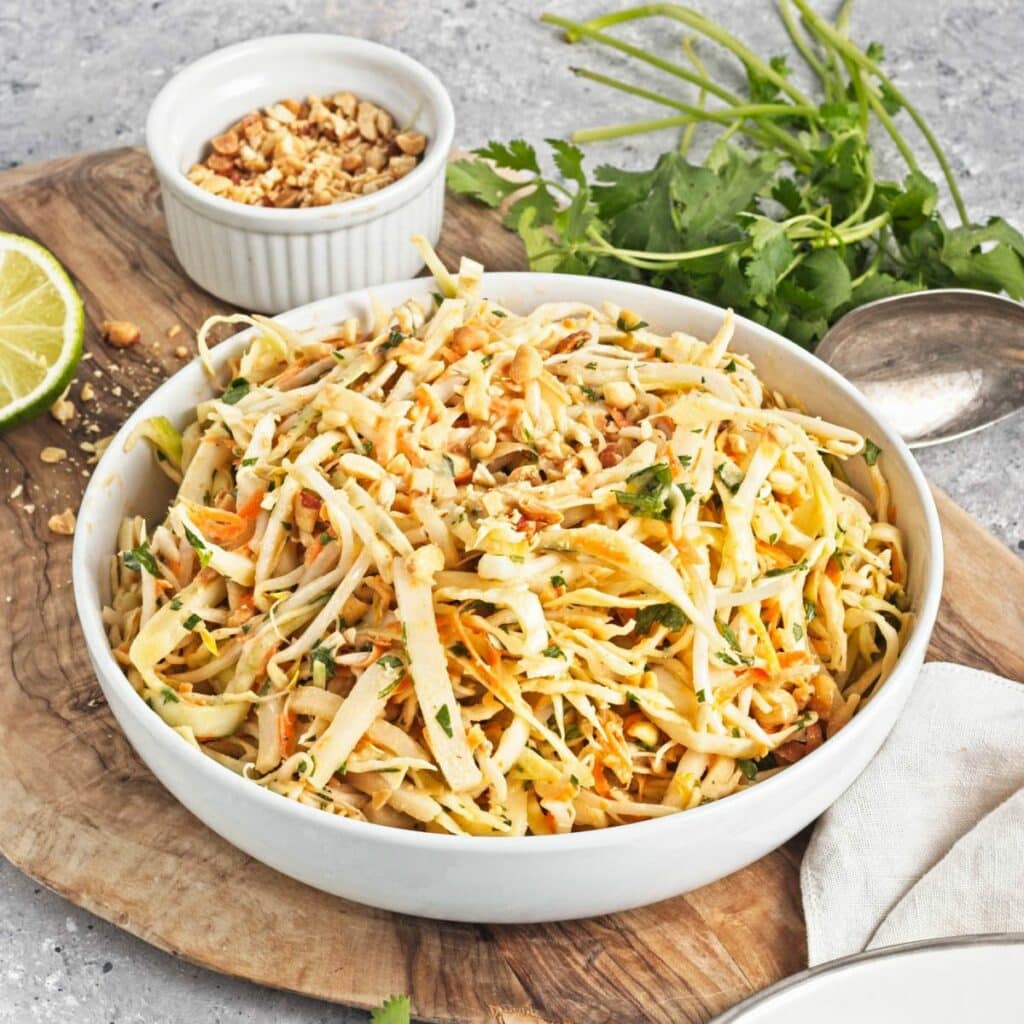 A white serving bowl and plate with shredded cabbage mix on a cutting board with limes, cliantro, and a bowl of peanuts.