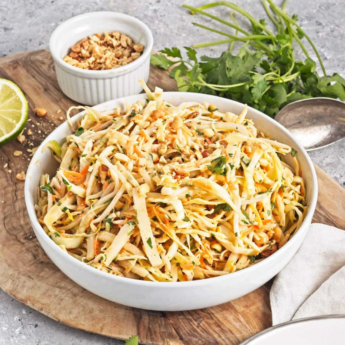 A white serving bowl and plate with shredded cabbage mix on a cutting board with limes, cliantro, and a bowl of peanuts.