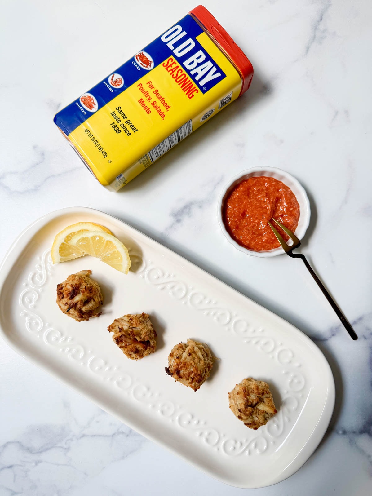 Crab balls on a white plate with lemon slices with a small bowl of cocktail sauce and a container of Old Bay seasoning.