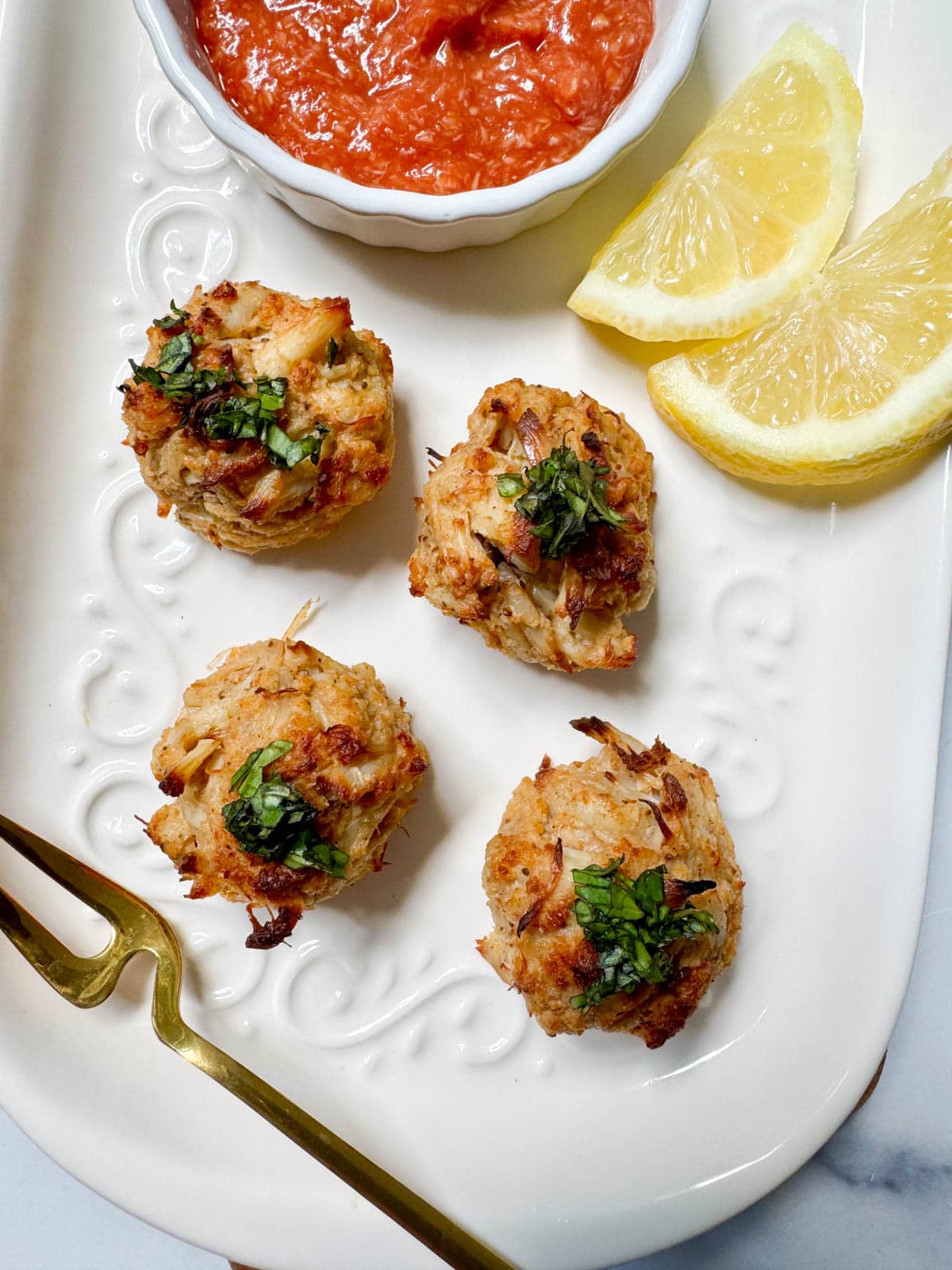 Crab balls on a white plate with lemon slices, a gold fork, and cocktail sauce in a white bowl.