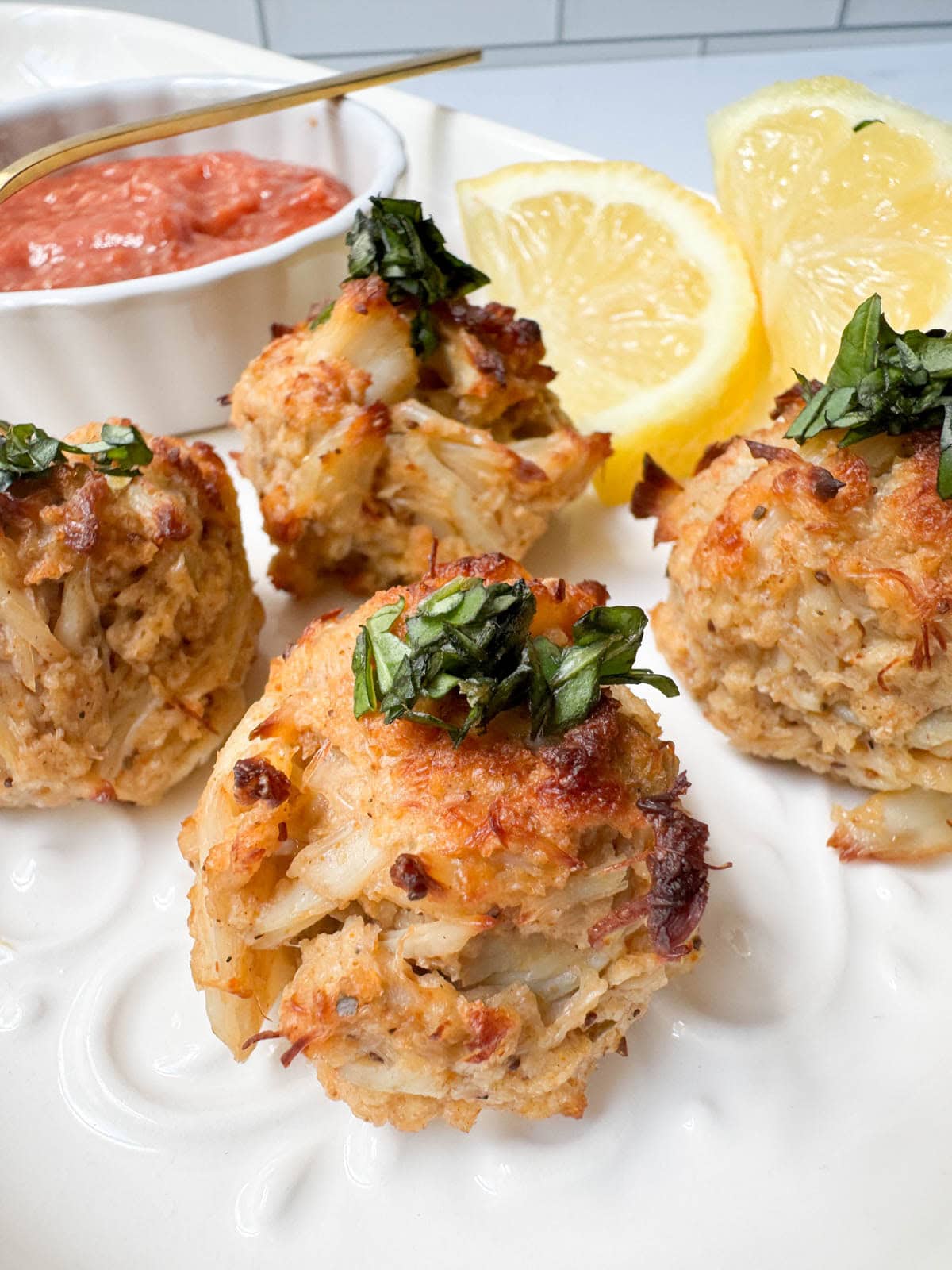 Close up of crab balls on white plate with lemon slices and cocktail sauce in a white bowl.