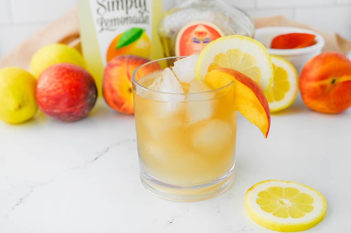 Golden colored cocktail with bottle of lemonade and bottle of Crown Royal in background with peaches and lemons.