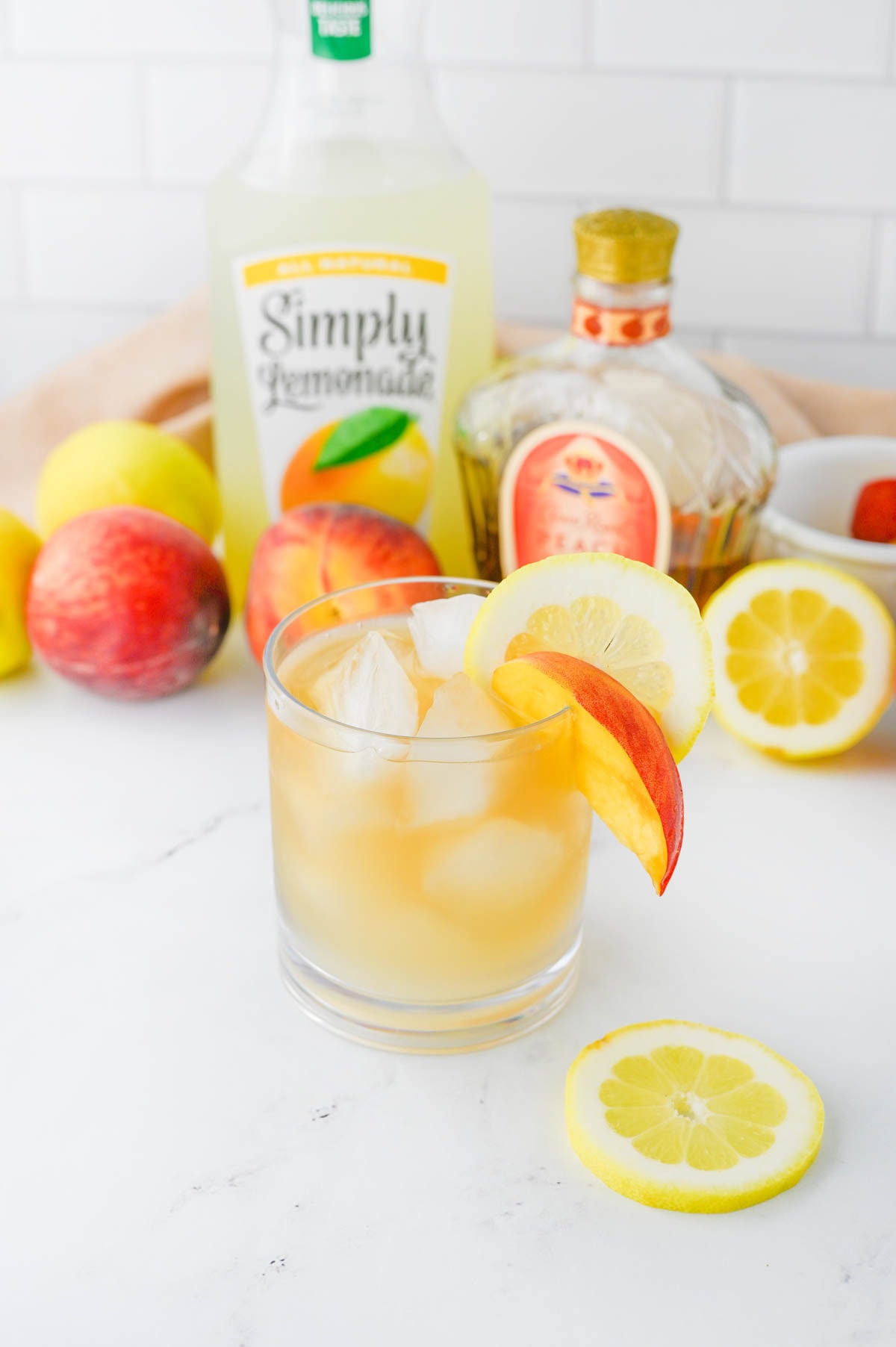 Golden colored cocktail with bottle of lemonade and bottle of Crown Royal in background with peaches and lemons.