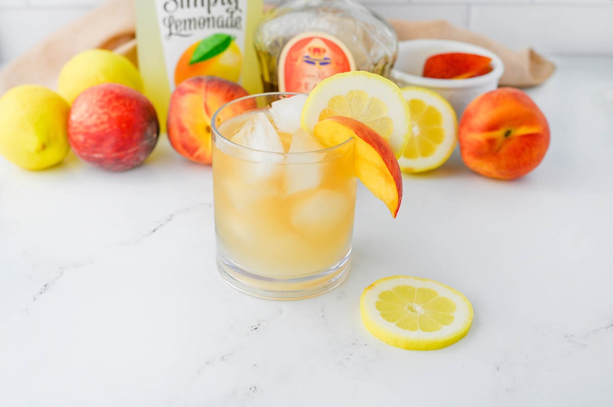 Golden colored cocktail with bottle of lemonade and bottle of Crown Royal in background with peaches and lemons.