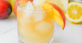 Golden colored cocktail with bottle of lemonade and bottle of Crown Royal in background with peaches and lemons for Pinterest graphic.