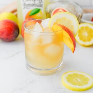 Golden colored cocktail with bottle of lemonade and bottle of Crown Royal in background with peaches and lemons.